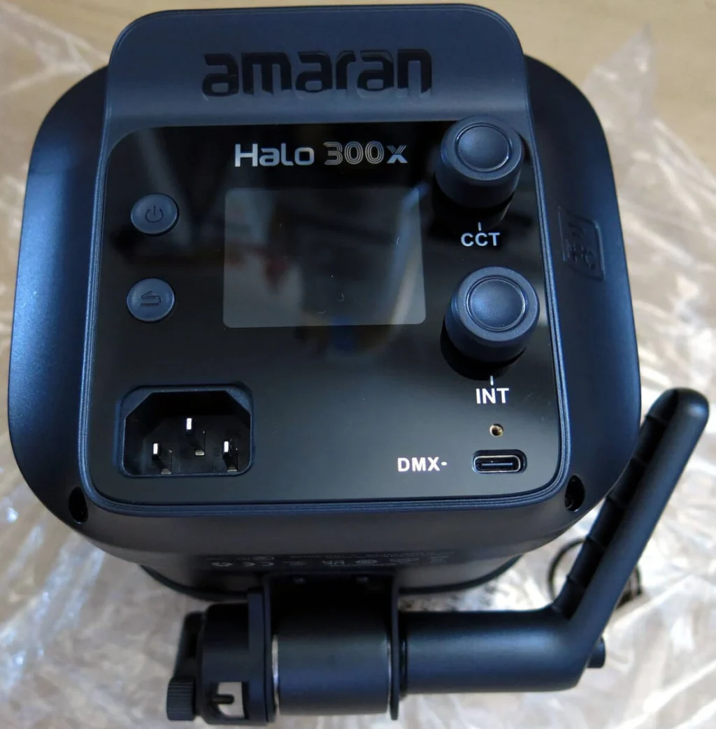 Вигляд згори на задню панель керування Amaran Halo 300x: логотип amaran, вимкнений LCD-екран з відблиском, поворотні ручки CCT та INT, роз'єм живлення AC, порт DMX і USB-C; прилад ще в захисній плівці, закріплений на кронштейні стійки.